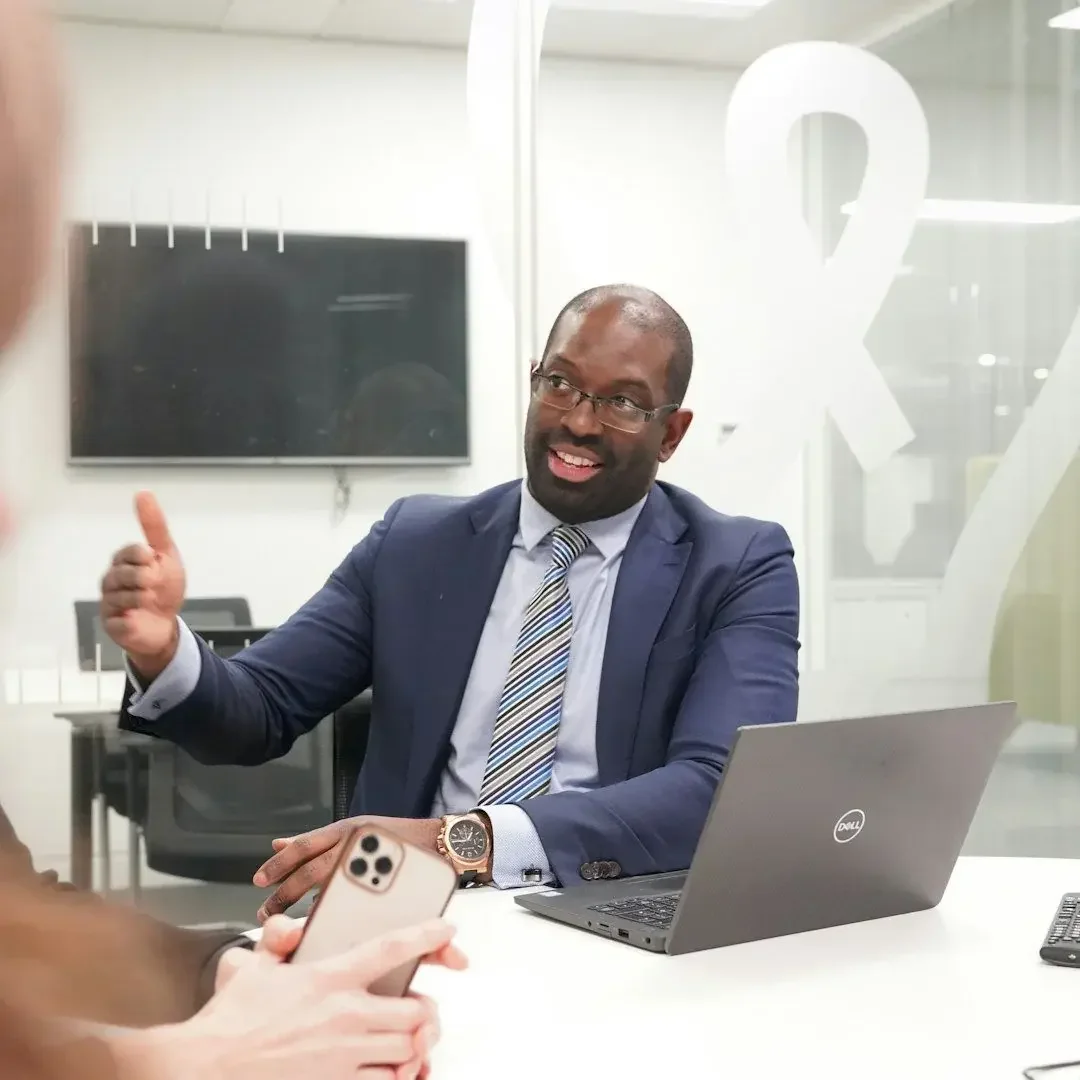 Man in a suit sitting at a conference table, talking and gesturing with a person holding a phone, with a laptop and a TV monitor in the background.