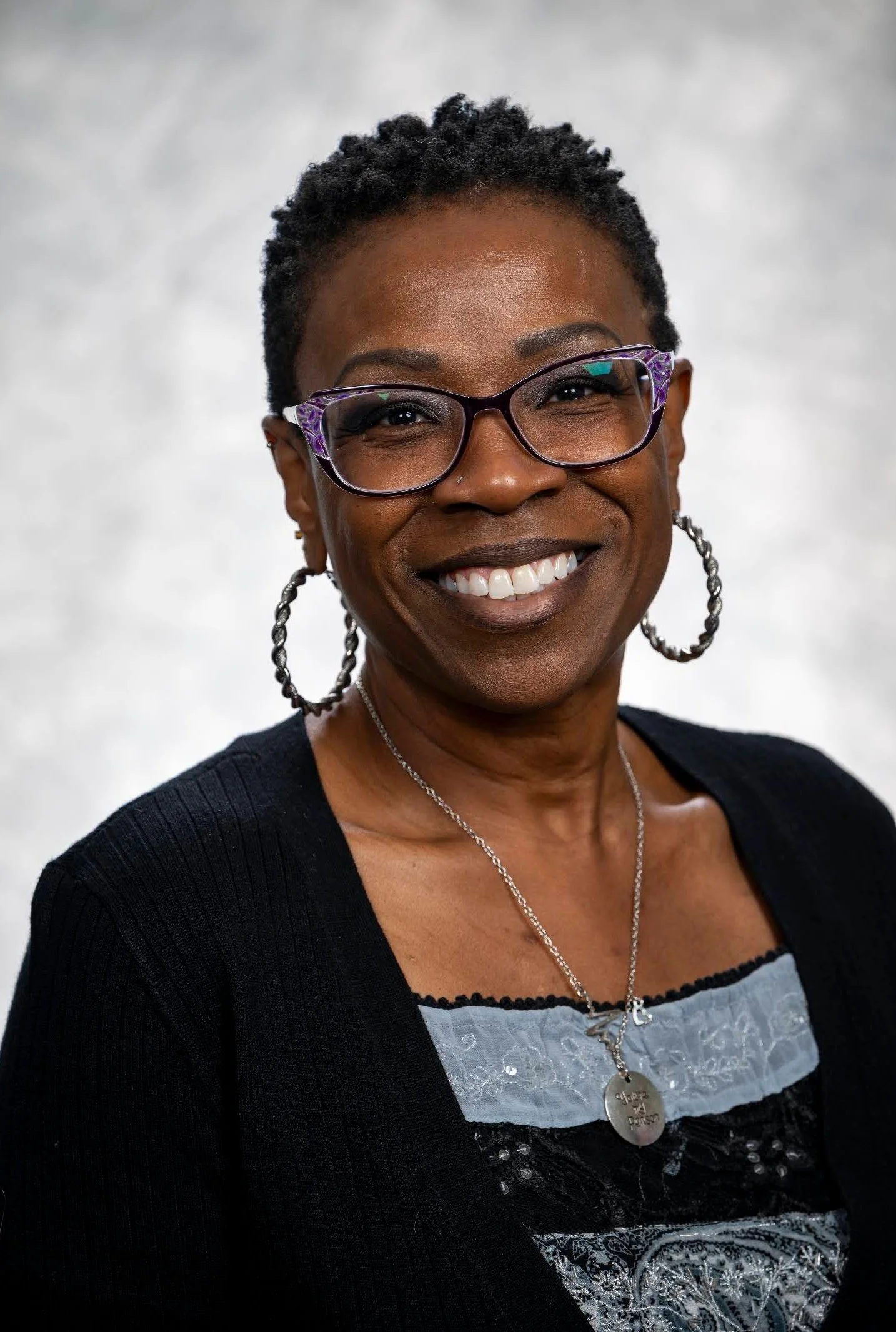 Close-up of a smiling African American woman wearing glasses with purple frames, hoop earrings, a necklace with a pendant, and a black cardigan over a patterned blouse, against a plain background.