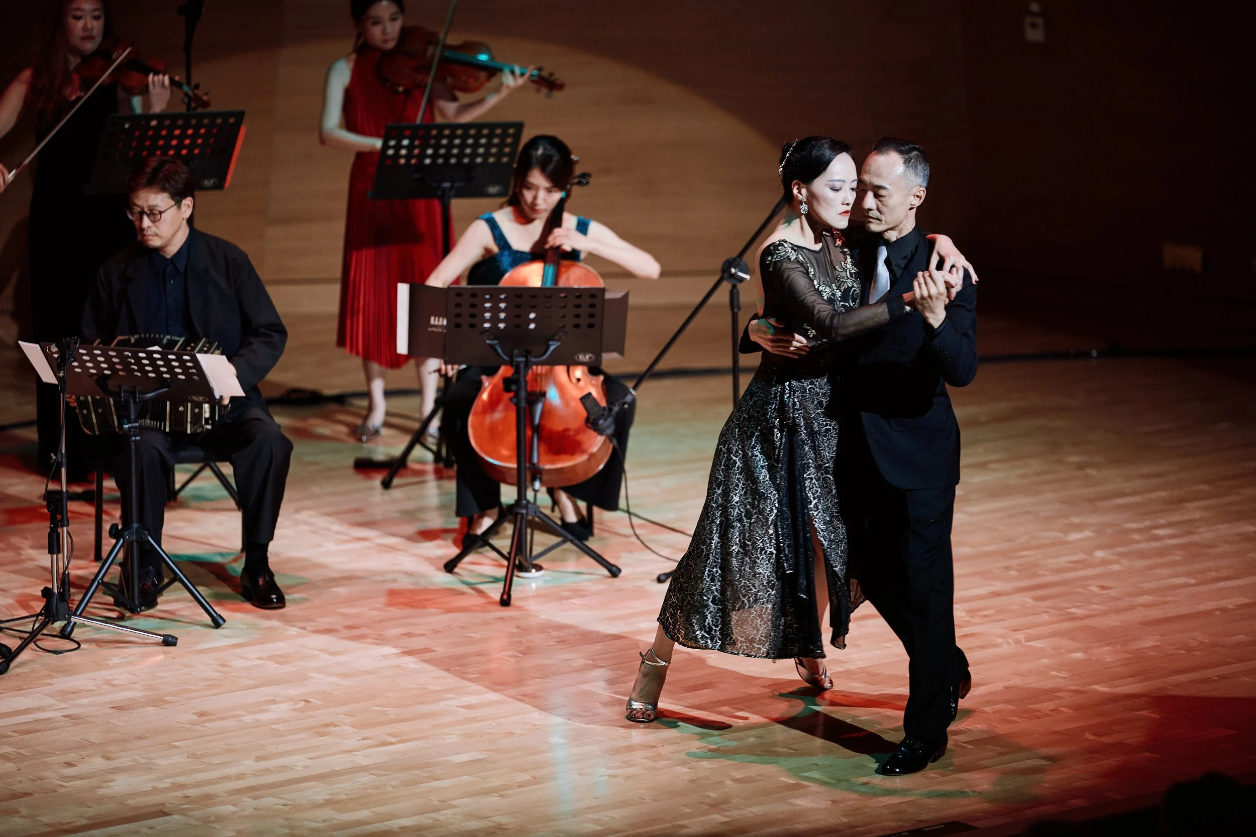 A couple dancing passionately on stage, with musicians playing string instruments and a keyboard, in a dimly lit performance hall.