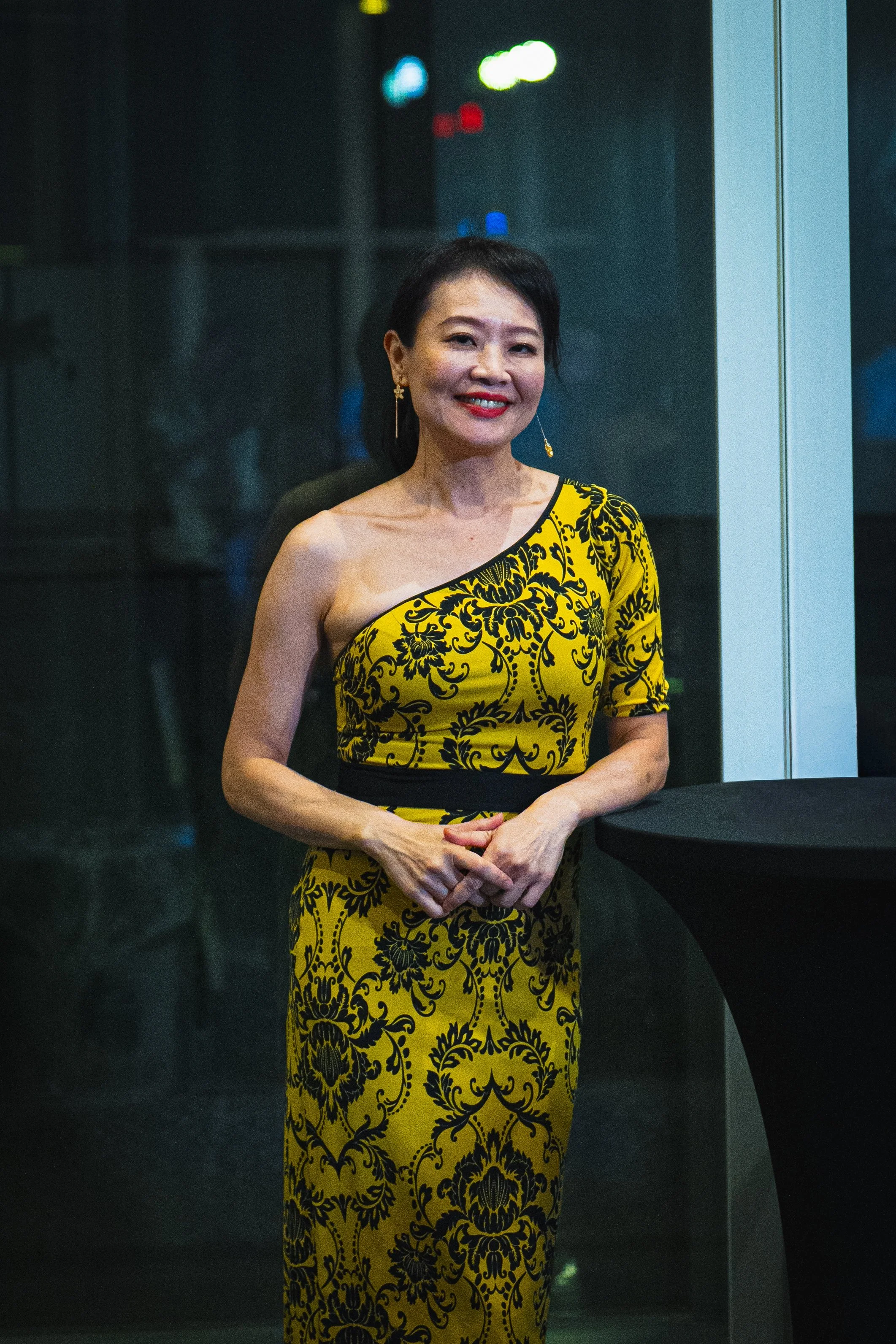 A smiling woman standing indoors in front of a glass wall with blurred lights behind her. She is wearing a yellow and black patterned off-shoulder dress and gold earrings.