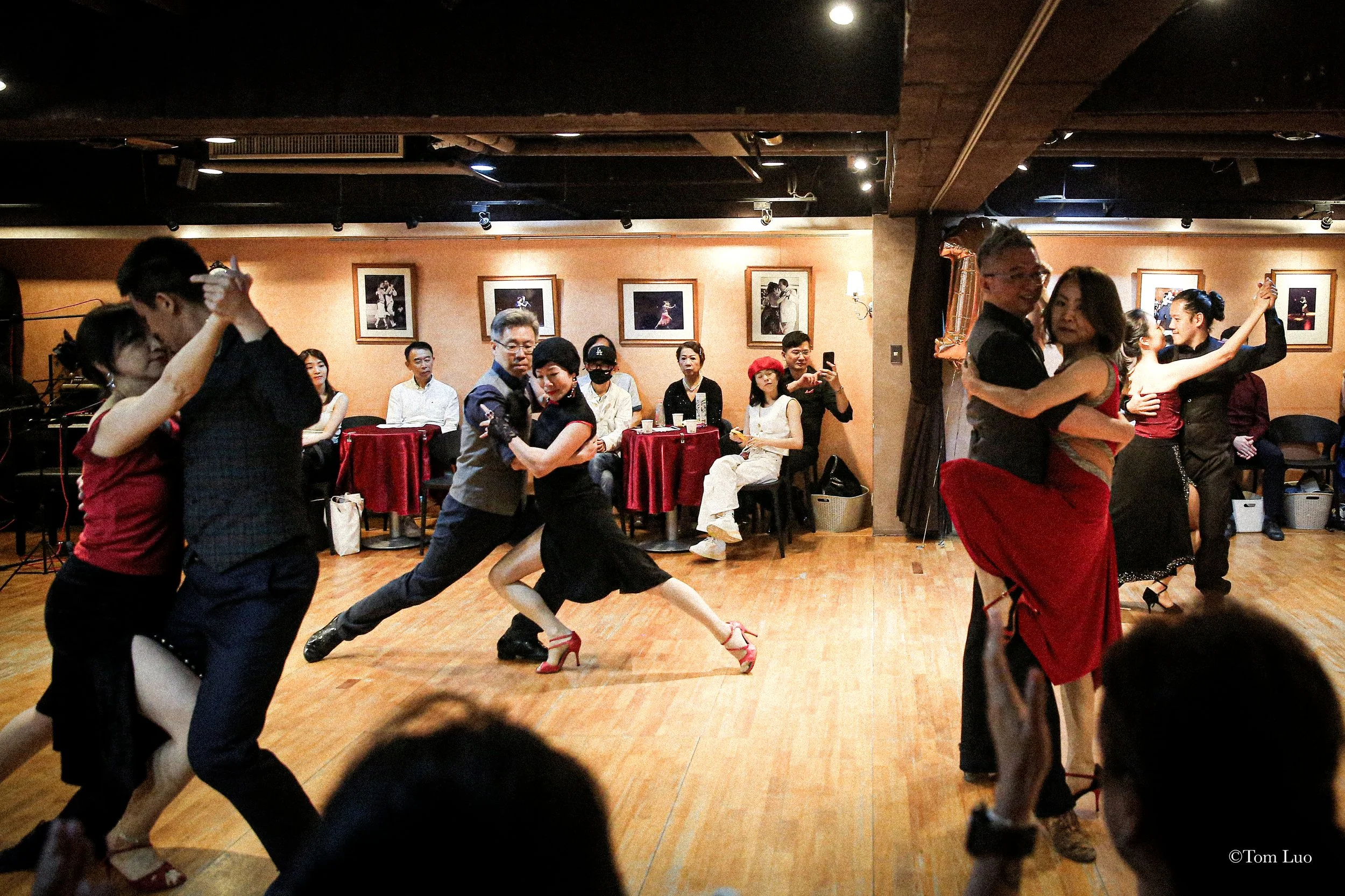 Couples dancing in a ballroom with a wooden floor, surrounded by seated onlookers and framed photographs on the walls.