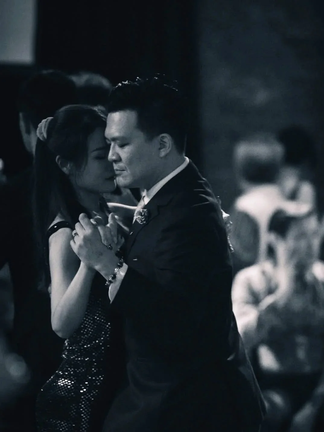 A black-and-white photo of a man and woman dancing closely at a formal event, with other people in the background.