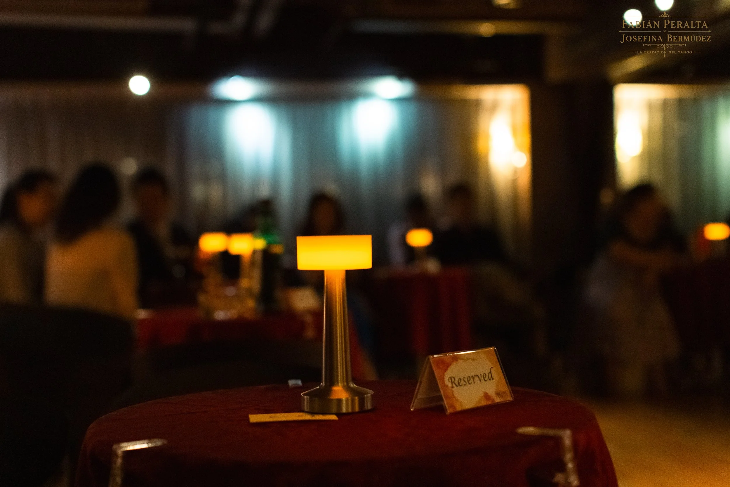 A dimly lit dining area with a small table illuminated by a warm, orange table lamp. In the background, blurred figures of people dining and a sign indicating a reserved table can be seen. The ambiance appears cozy and intimate.