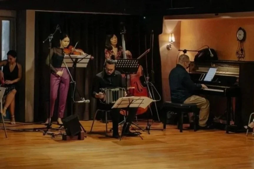 Musicians performing on stage, including a pianist, violinist, double bassist, and a musician with an accordion, in a cozy, dimly-lit venue with hardwood floors.