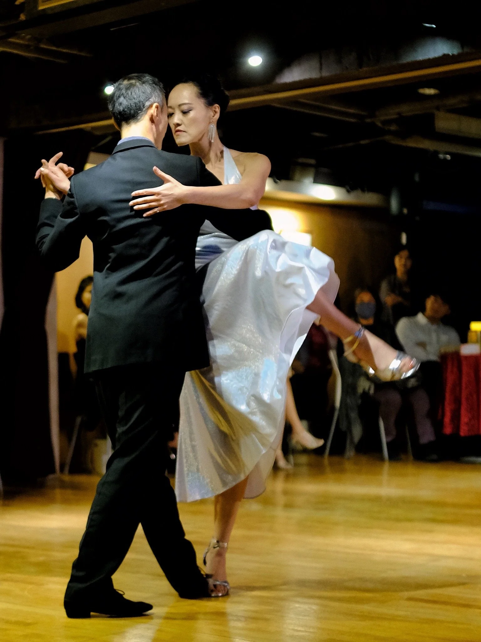 A man and a woman are dancing closely in a ballroom, with the woman lifted off the ground by the man. The woman is wearing a white dress and high heels, while the man is dressed in a dark suit. There are several onlookers in the background seated and observing the dance.