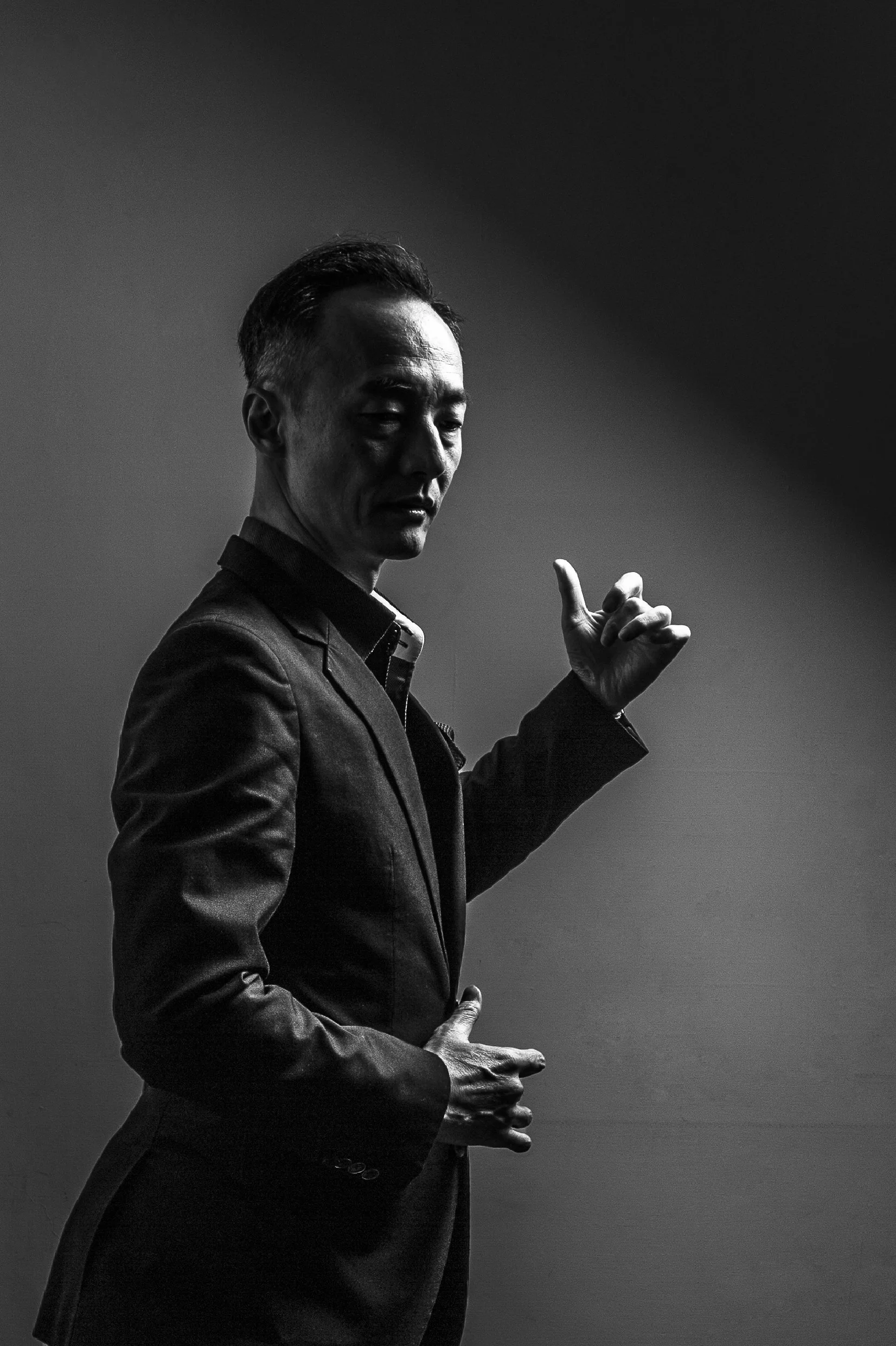 Black and white photo of a man in a suit with styled hair, pointing upward with his right index finger, against a plain background.