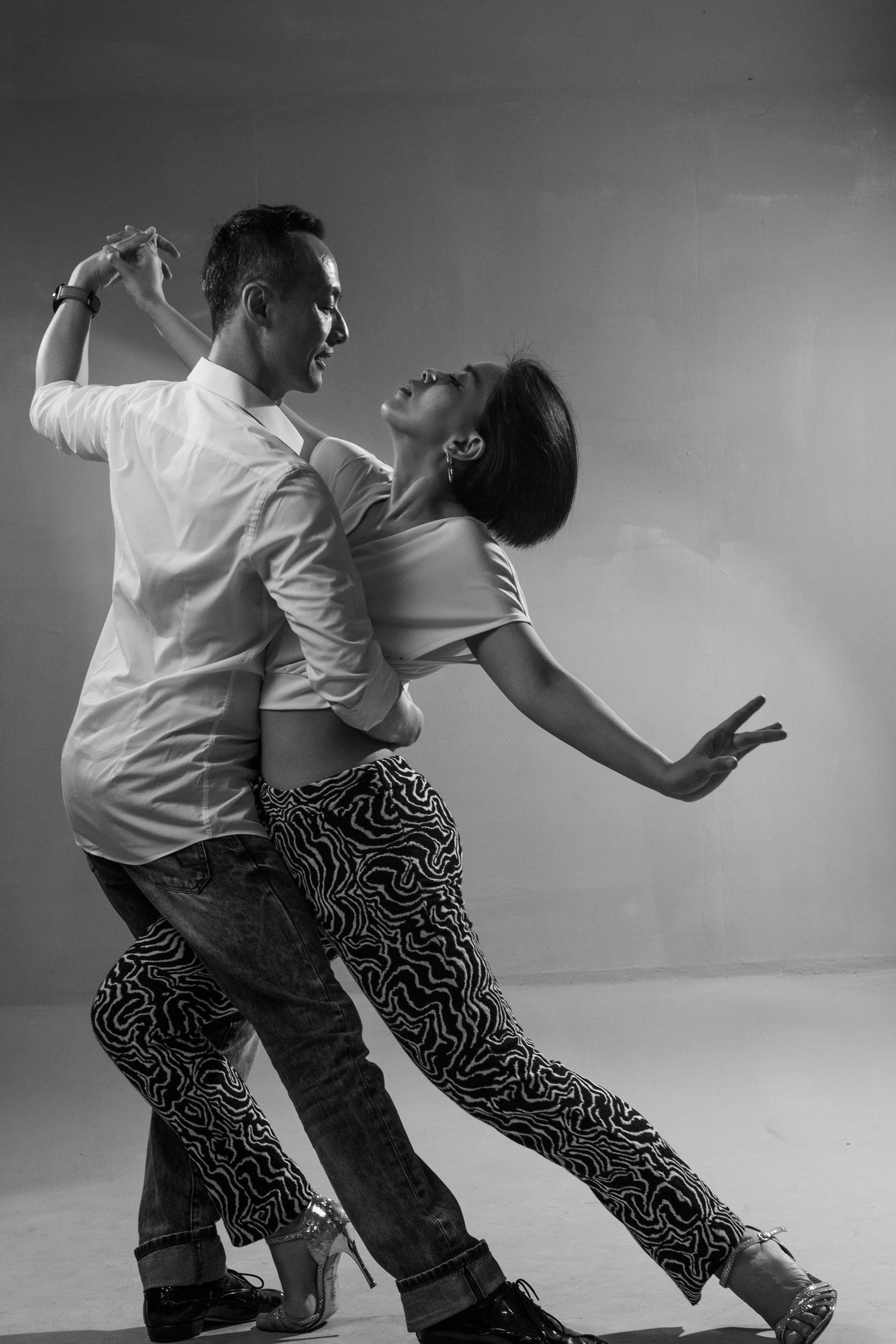 A black and white photo of a dancing couple, the woman is wearing zebra print pants and high heels, with her arm extended, and the man is wearing casual jeans and a white shirt, with his arms around her, both looking at each other.