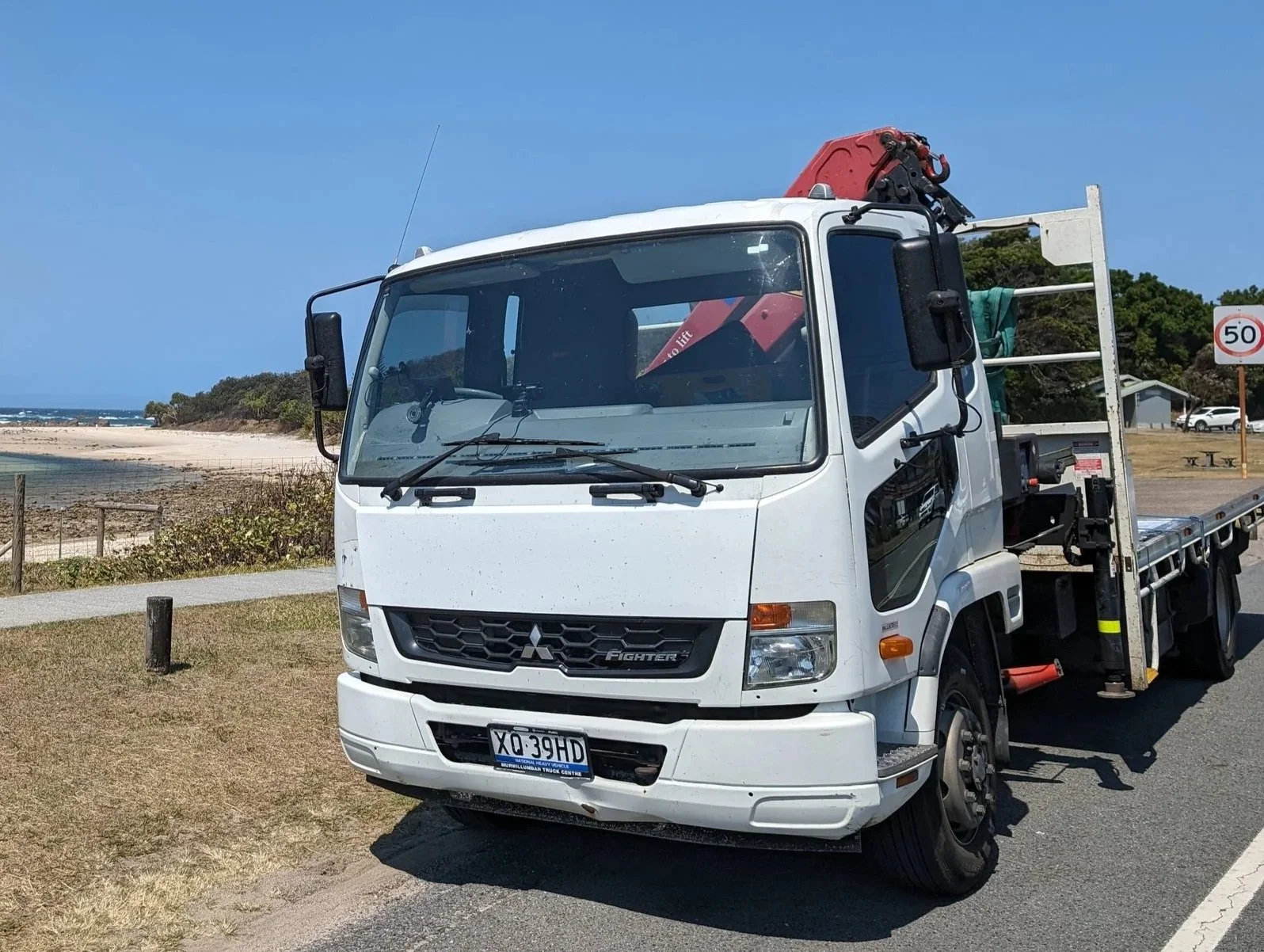 White crane truck parked on the side of a road near a beach with trees.