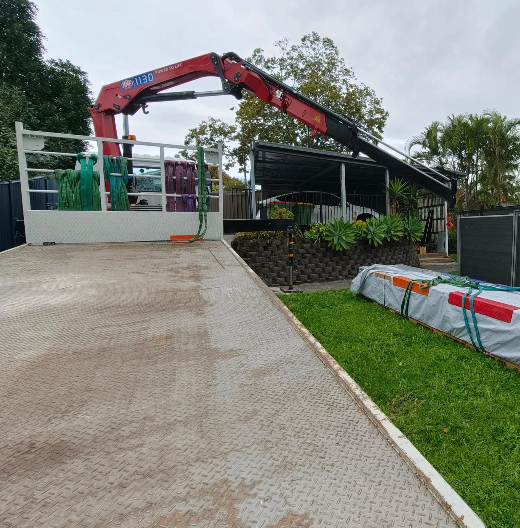 A flatbed truck with a red crane lifting a long, wrapped object, with some plants and cars in a residential area.
