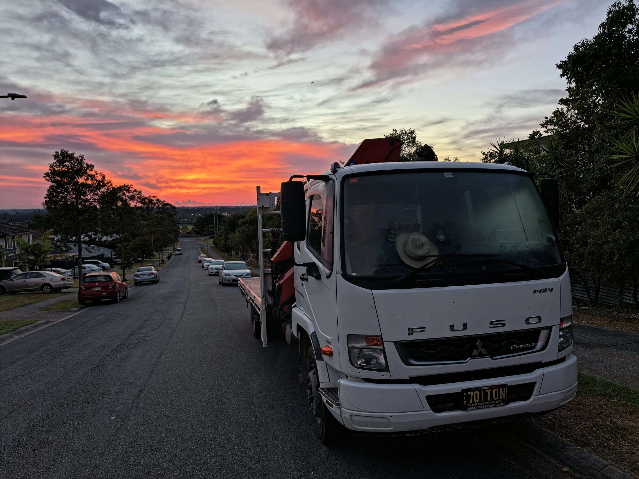 A white crane truck parked on the side of a residential street during sunset with a colorful sky and clouds, trees, and houses in the background.