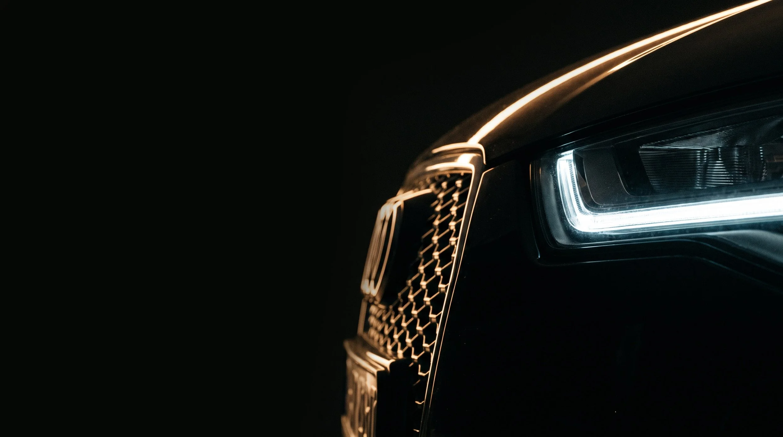 Close-up of the front of a luxury black car, showing the intricate grille, headlight, and sleek design, with a dark background.