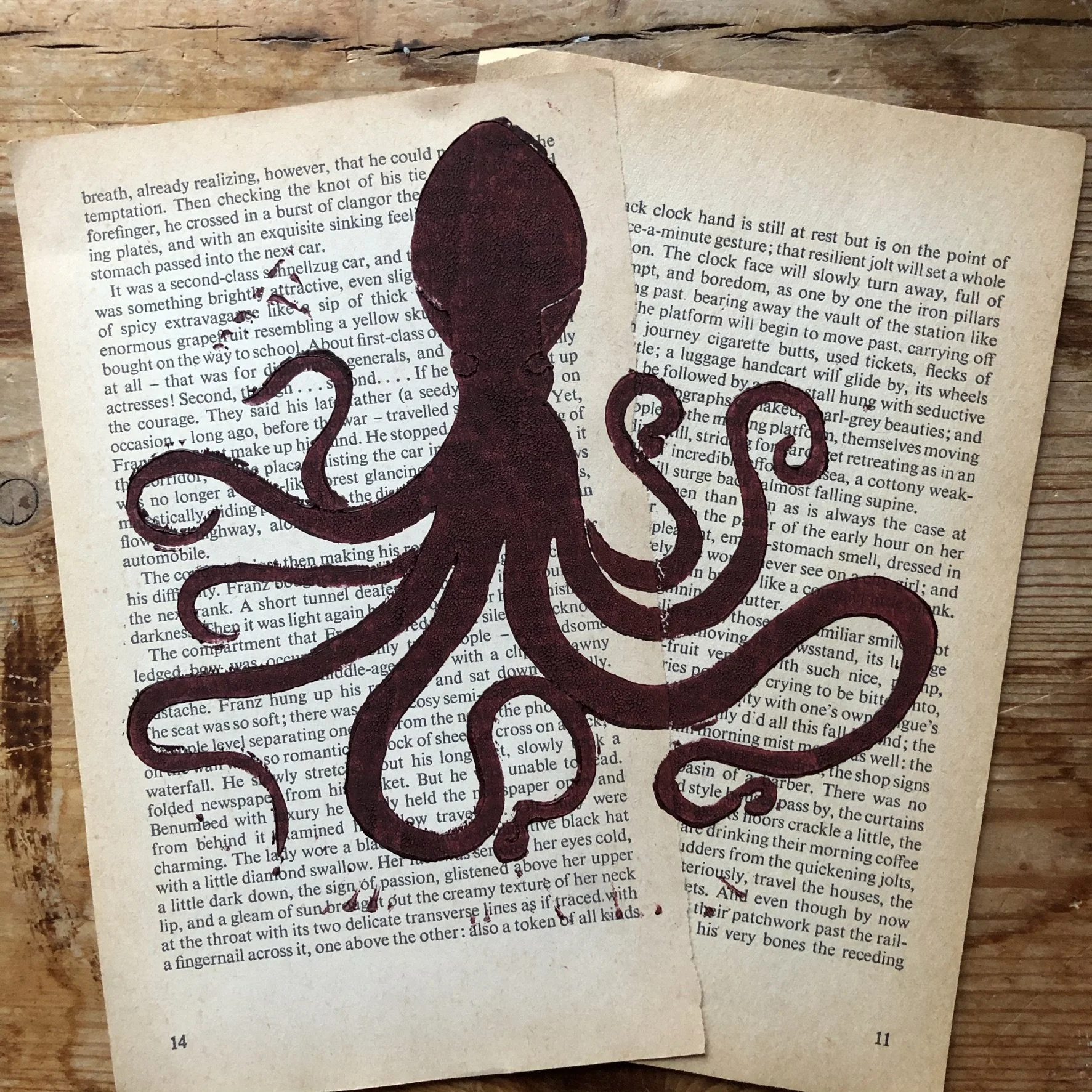 Photograph of an open book on a wooden surface with a dark purple octopus illustration painted over the pages. The octopus's head is centered on the pages, and its tentacles extend outward, covering some of the text.