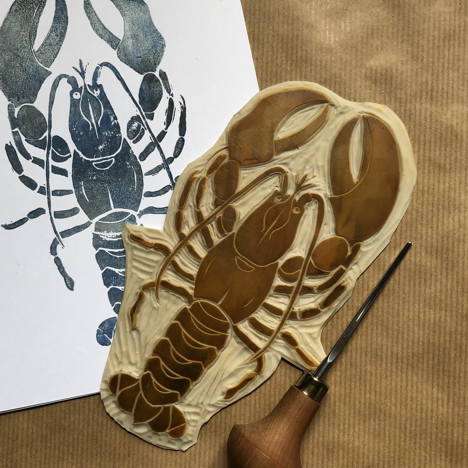 A black and white print of a lobster next to a brown and cream carved wooden lobster sculpture on a tan surface, with a tool also on the surface.