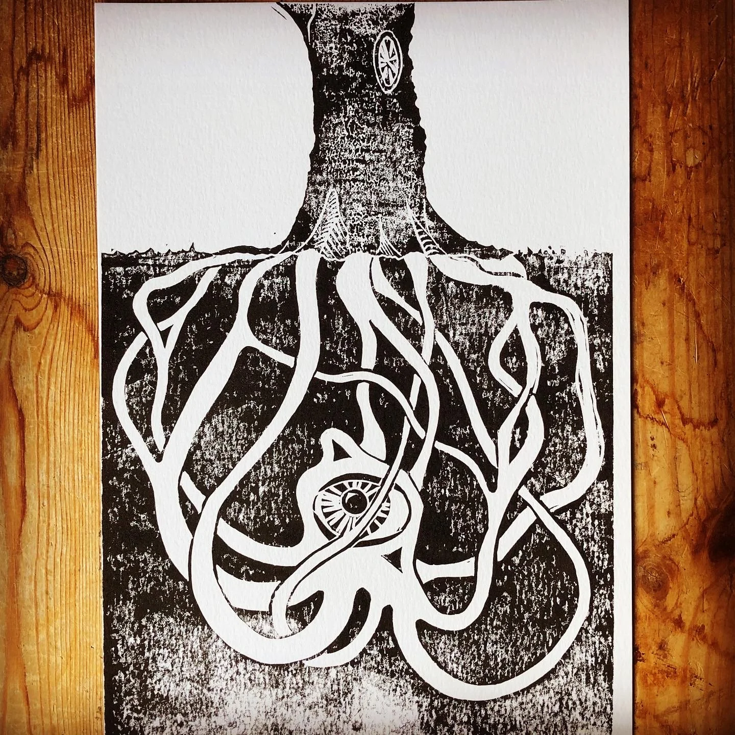 A black-and-white print depicting a tree with roots that extend and morph into various intertwined shapes and patterns. The tree has a circular emblem or symbol on its trunk. The print is on a wooden surface.