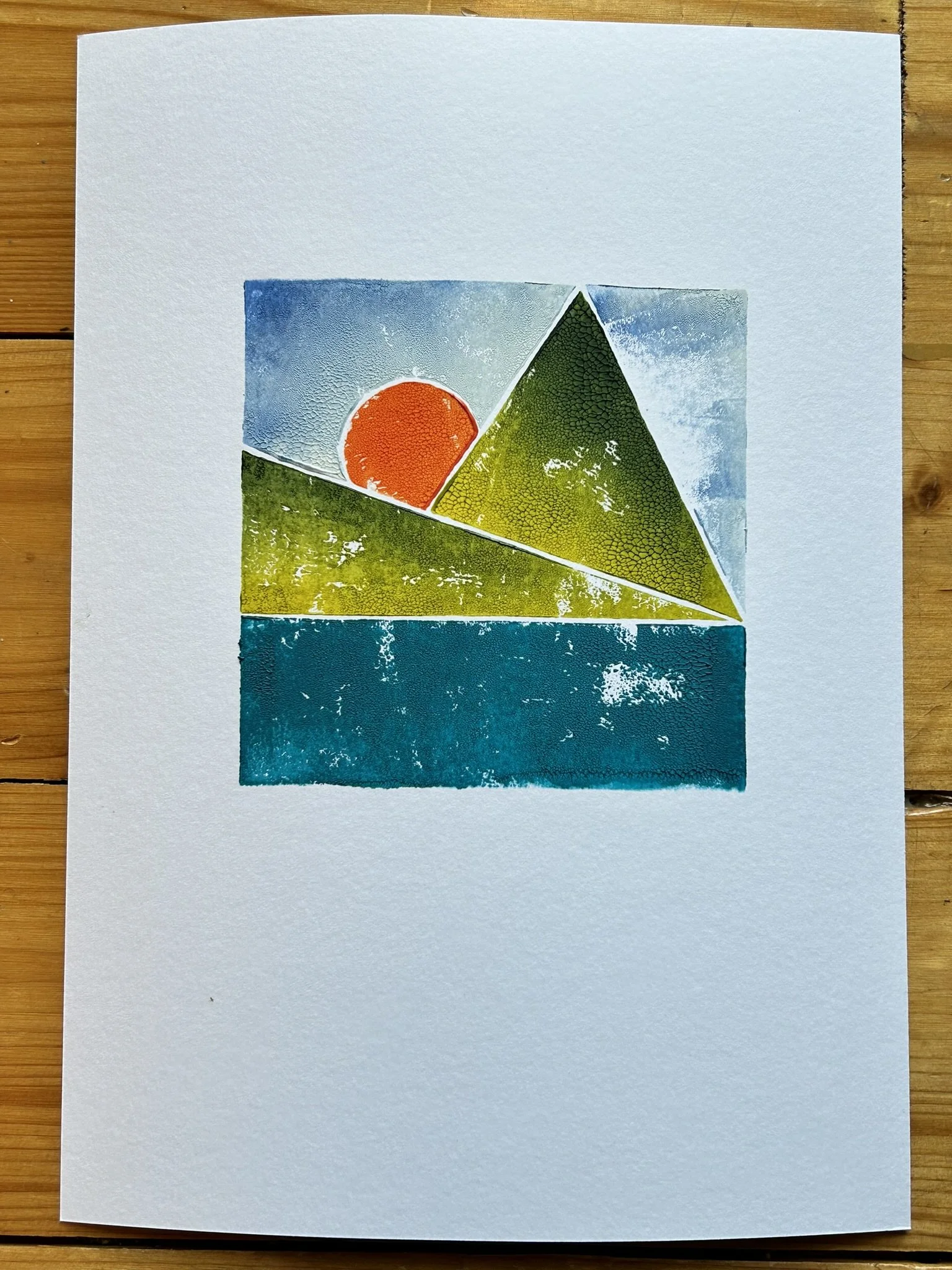 Linocut block print a landscape with a blue sky, a red sun, green mountains, and water at the bottom.