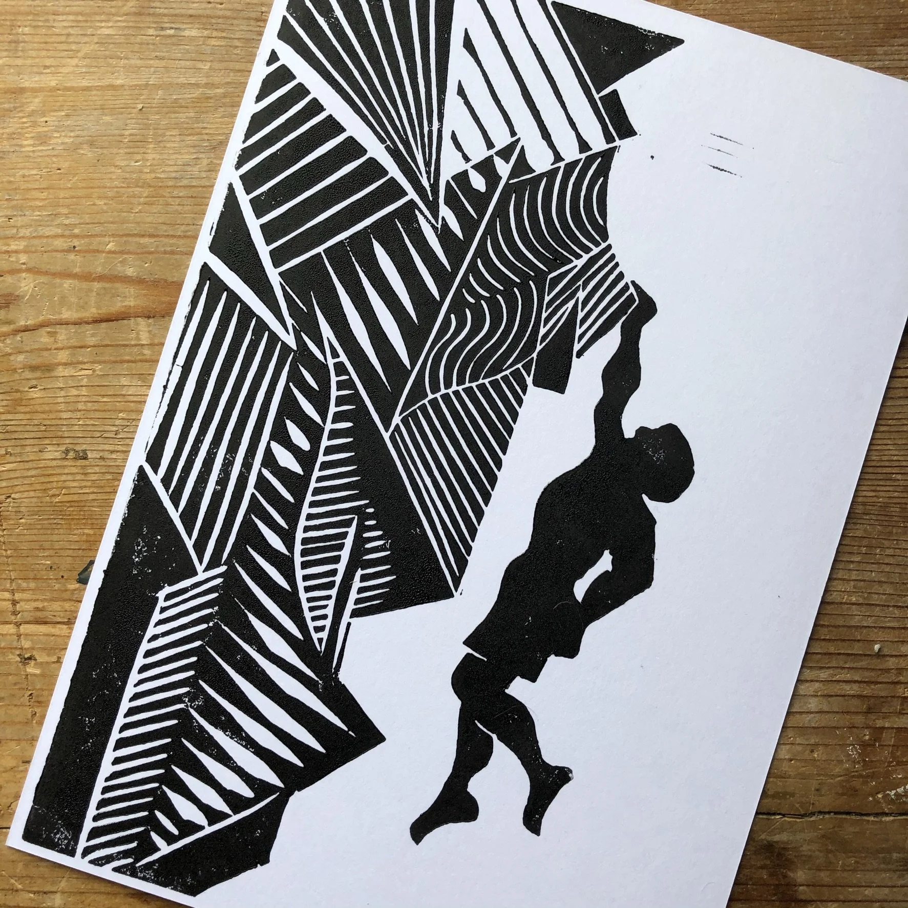 A black and white artistic print of a person holding a geometric patterned paper or sign, with a wooden surface in the background.
