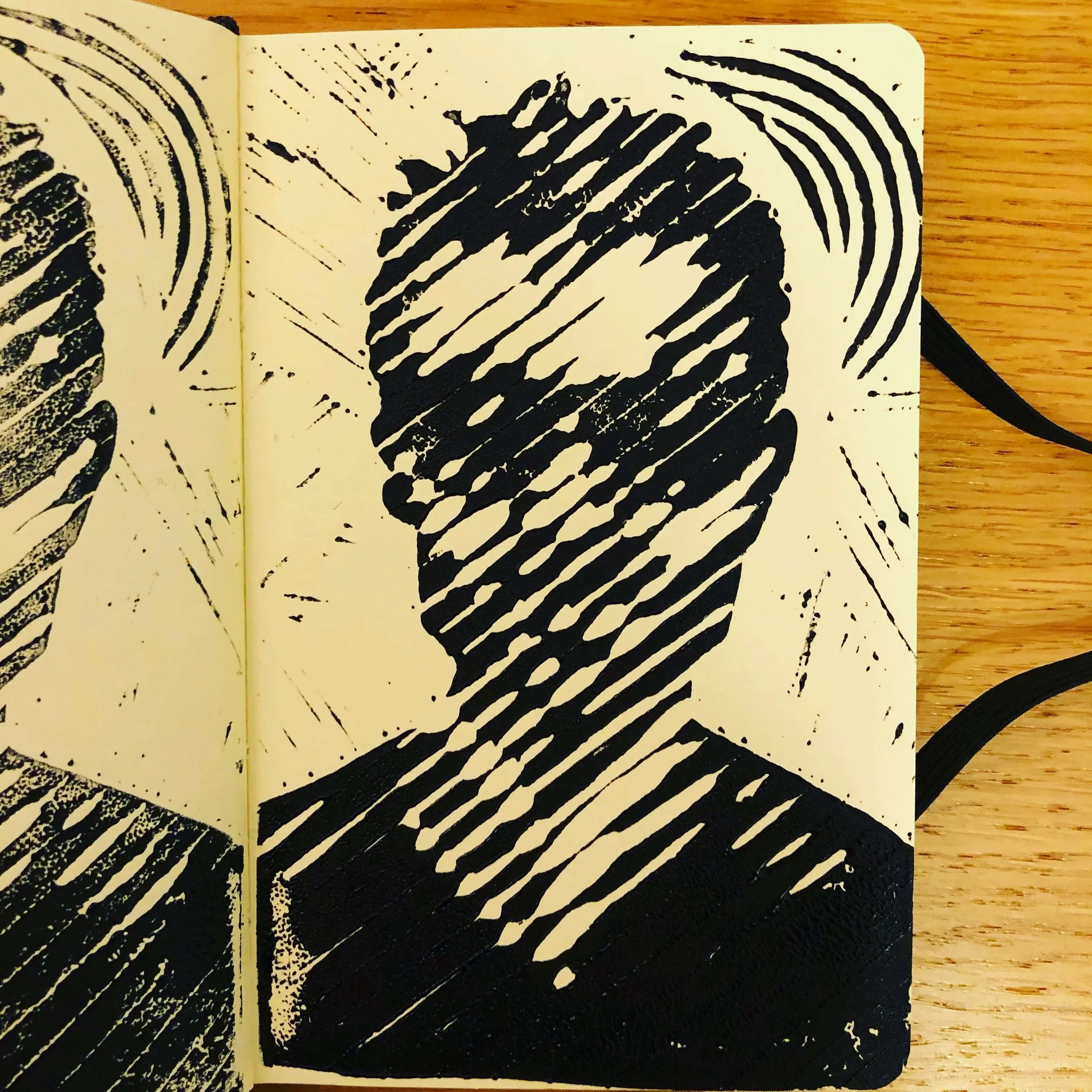 An open sketchbook with black linocut portrait design featuring diagonal lines, circles, and radiating patterns on cream-colored pages, resting on a wooden surface.