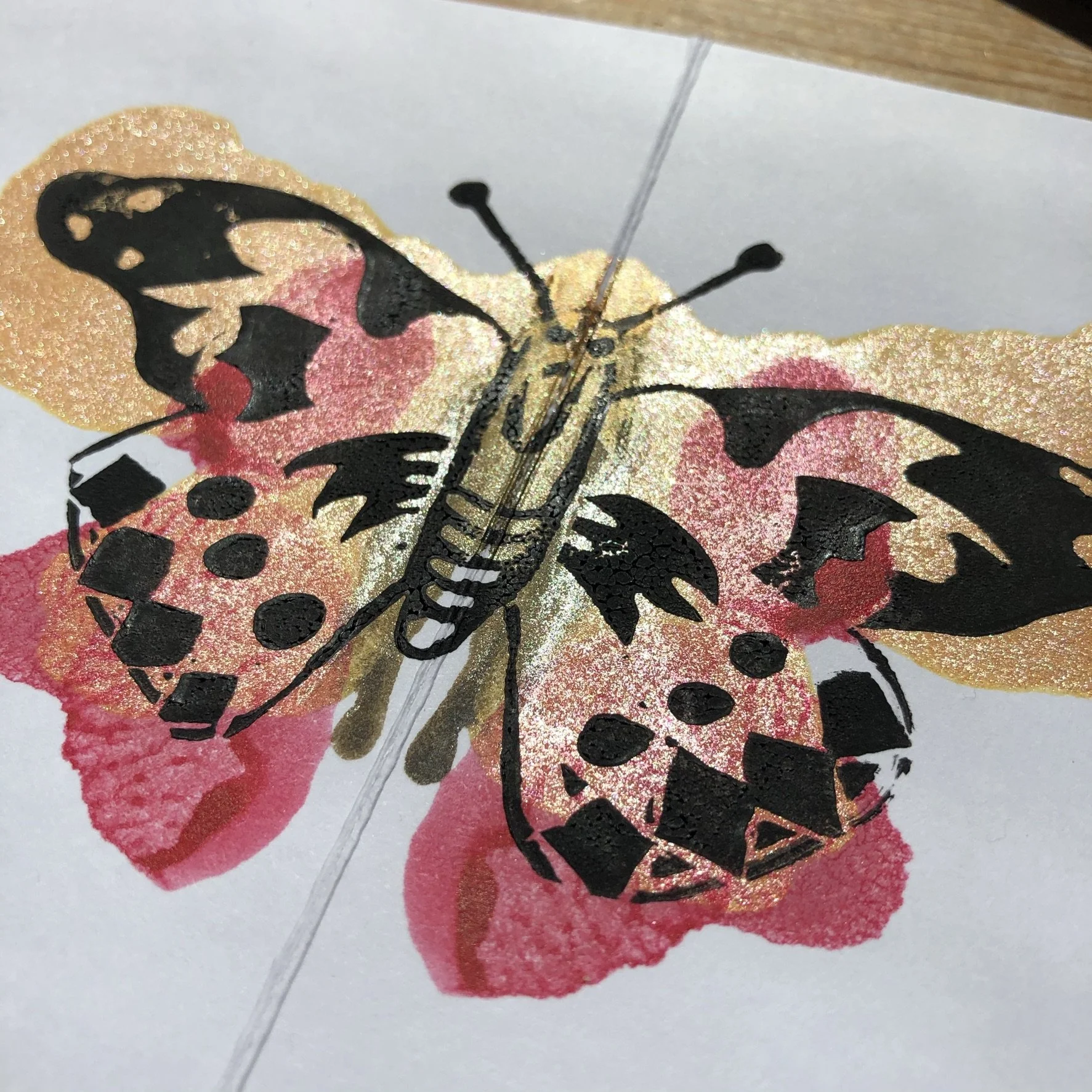 Close-up photo of a butterfly drawing with black and pink details, mounted on a white paper background with a faint diagonal fold, and a wooden surface edge at the top.