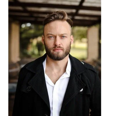 Portrait of a man with a beard and short hair, wearing a white shirt and black jacket, standing outdoors with trees and a structure in the background.