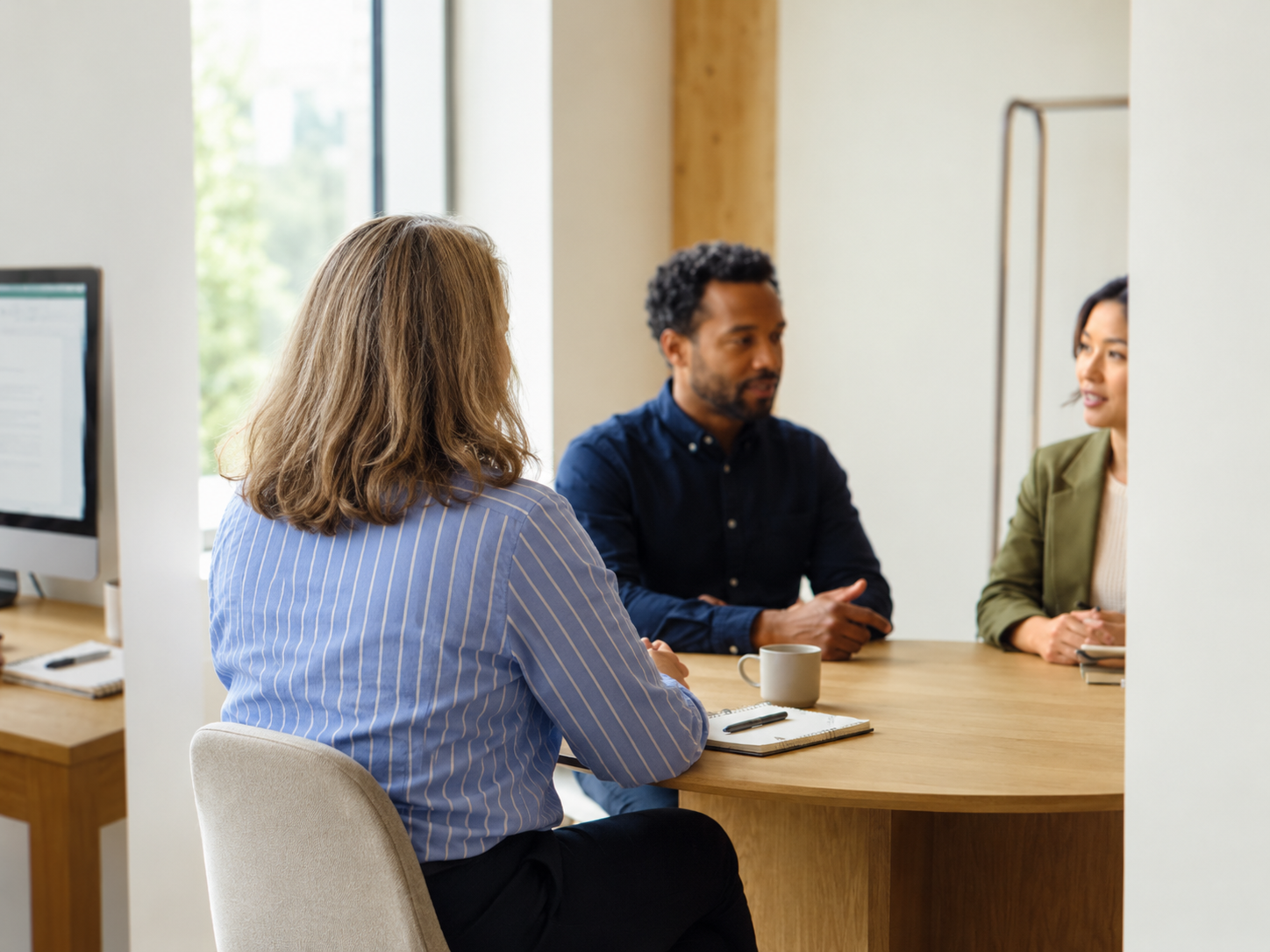Three people having a discussion in a modern office. Two women and one man sit at a wooden table, with a computer and notebook. Great planning advice and communication