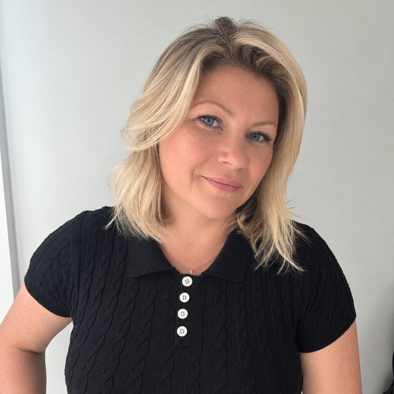 A woman with blonde hair and blue eyes wearing a black short-sleeve polo shirt with white buttons, smiling gently. Planning Consultant