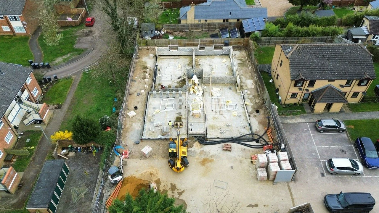 An aerial view of a construction site where a building is being built. The foundation is laid, and construction equipment, including an excavator, is present. Planning permission secured, approved, granted, achieved