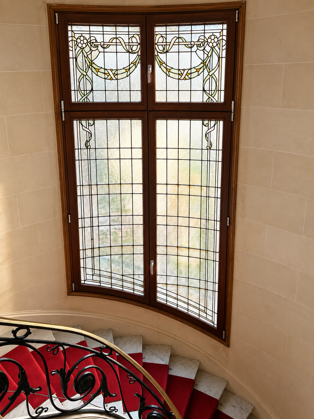 Fenêtre en verre coloré avec motif floral dans un mur beige, vue depuis un escalier en colimaçon avec rampe en fer forgé noire et tapis rouge.