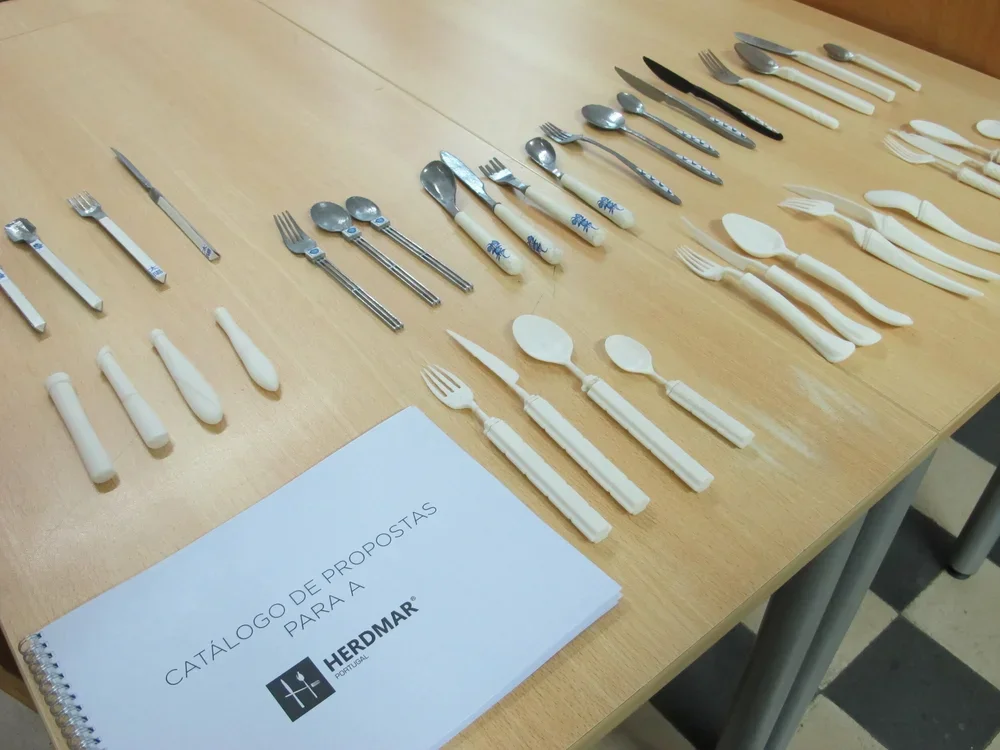 Various types of cutlery including knives, forks, and spoons, arranged on a wooden table alongside a catalog booklet titled "Catálogo de Propostas para A HERDMAR."