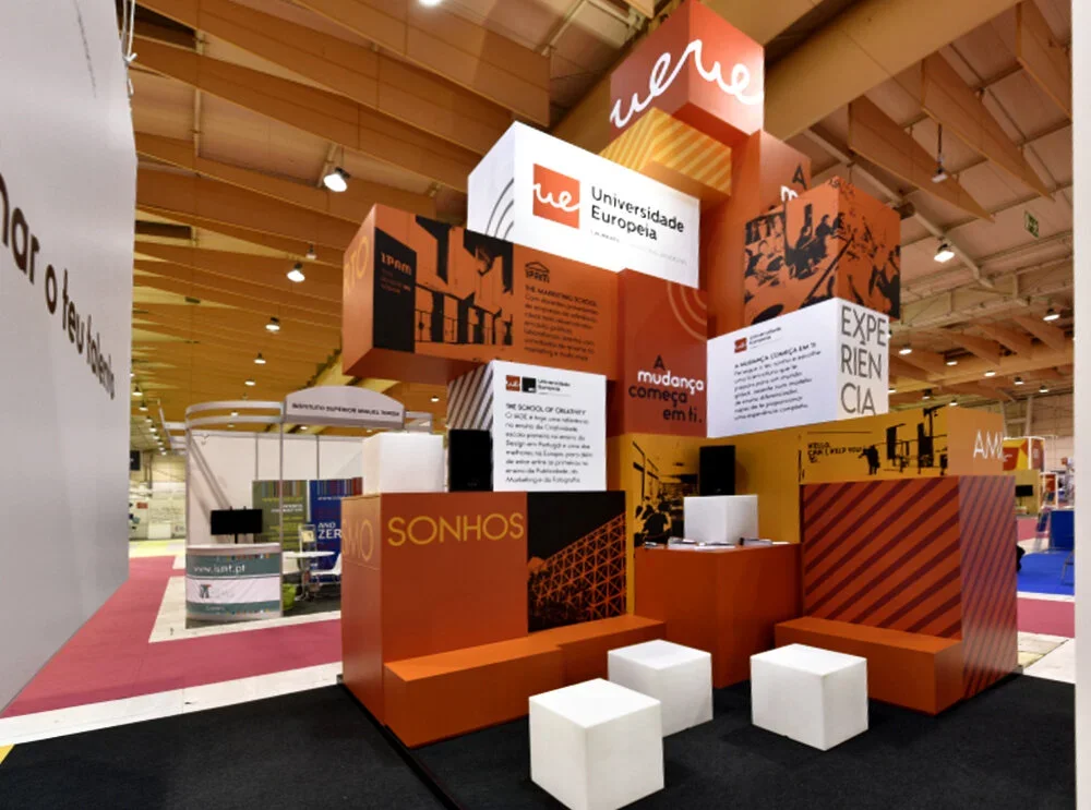 Exhibition booth with branding for Universidade Europeia, featuring orange, black, and white design elements, informational panels, and seating cubes.