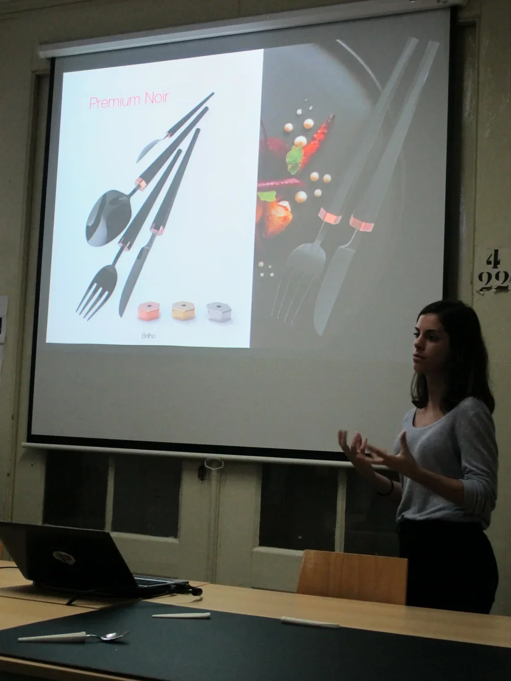 A woman gives a presentation on cutlery, showing a slide with a set of black utensils labeled 'Premium Noir' and three colored containers, with a background image of plated food and utensils.