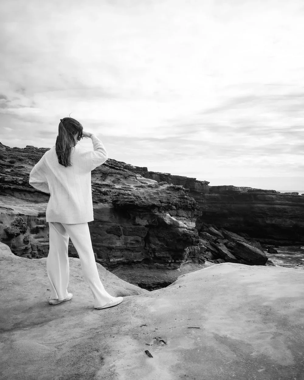 A woman with long hair, wearing a sweater and pants, stands on a rocky cliffside looking out at the ocean, with a cloudy sky above.