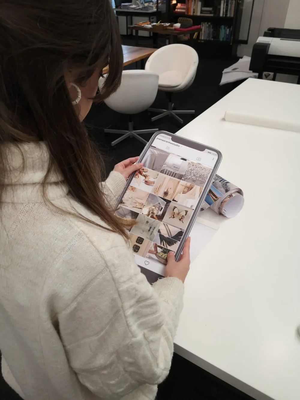 A woman with brown hair and earrings looking at her phone in a design or office space, with books and papers on shelves and tables, and a rolled-up magazine on the white table in front of her.