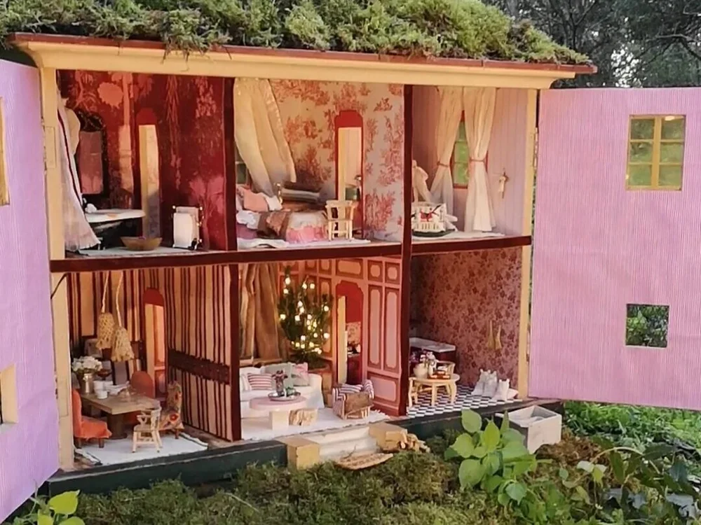 Detailed miniature dollhouse with two floors, decorated as a cozy living space with furniture, beds, a Christmas tree, and dining area, set outdoors with greenery around.