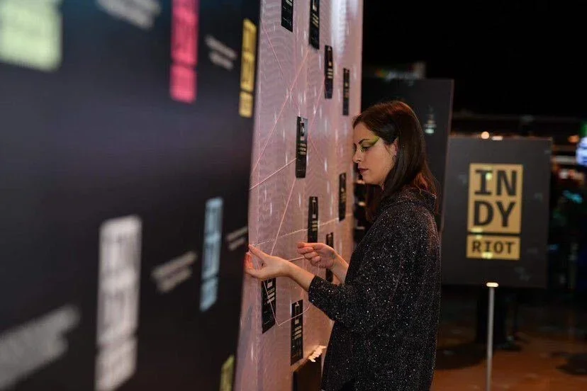 A woman with glasses and dark hair looking at an exhibit in a dimly lit indoor setting, with a sign reading 'INDY RIOT' in the background.