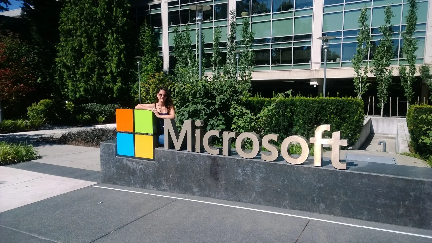 A woman in sunglasses leaning on a large Microsoft sign outdoors with greenery and modern glass building in the background.