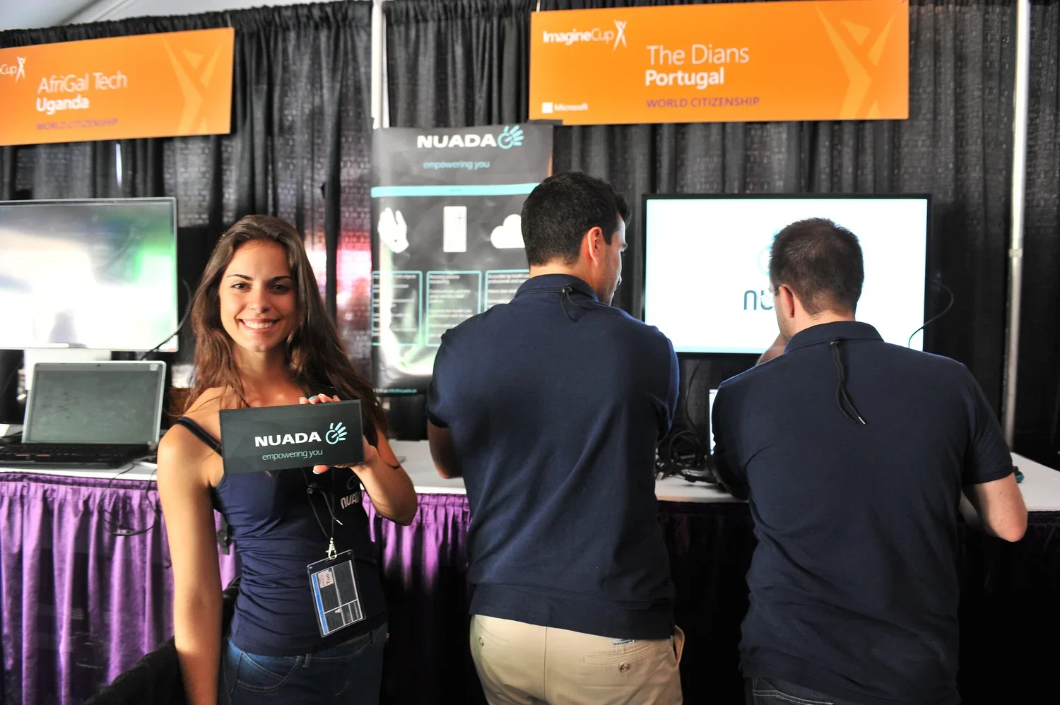 A woman smiling and holding a NUADA sign at a technology booth with two men working on computers behind her at a conference.
