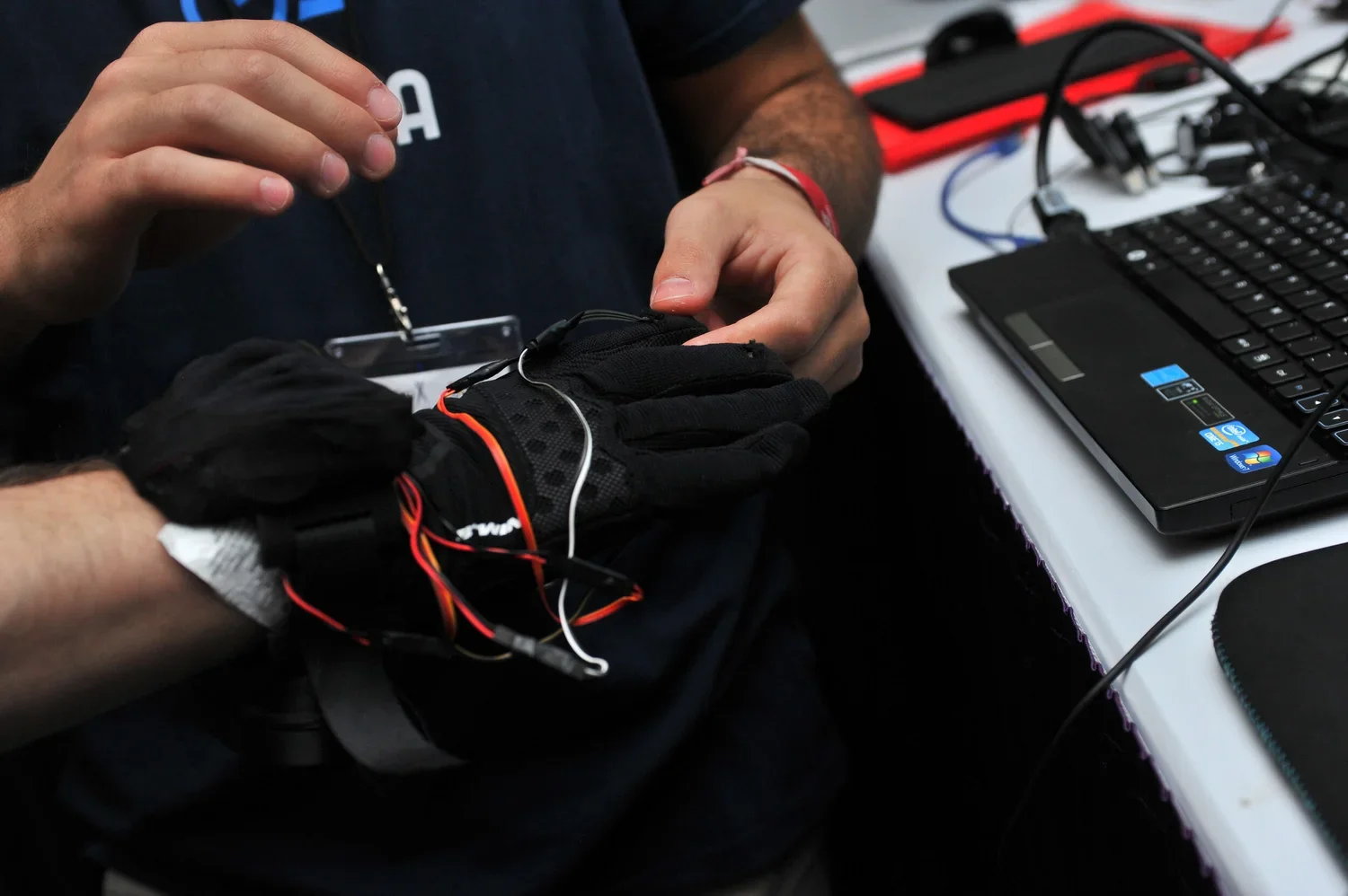 Person attaching electronic wires to a black glove with sensors during a technology demonstration, with a laptop and electronic equipment on the table.