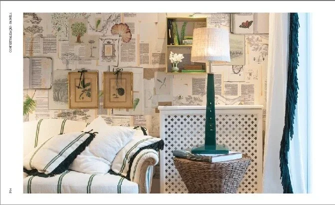 Living room corner with a white couch with black-striped pillows, a tall green table lamp, books, a wicker side table, and a wall decorated with botanical and vintage-inspired artwork and newspaper clippings.