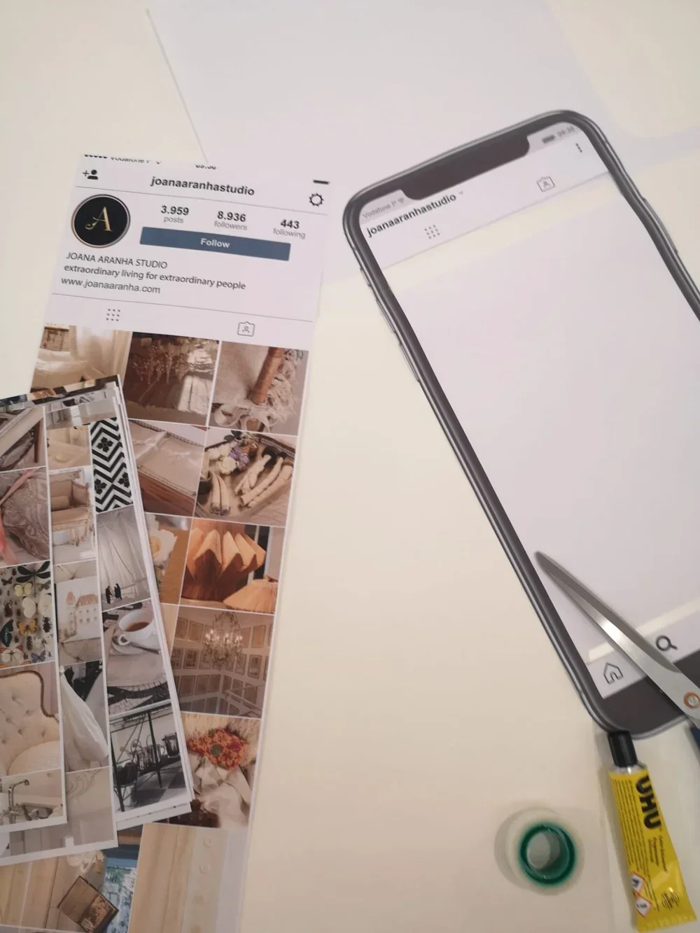 A table with a screen showing an Instagram profile of Joana Aranha Studio, a phone with a blank Instagram page, printed Instagram posts of home decor, a pair of scissors, a roll of scotch tape, and a tube of glue.