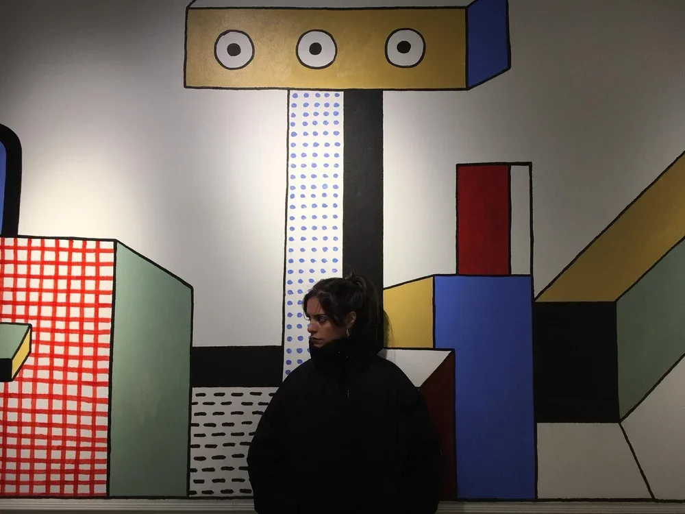 A woman standing in front of a colorful, abstract geometric mural on a wall.