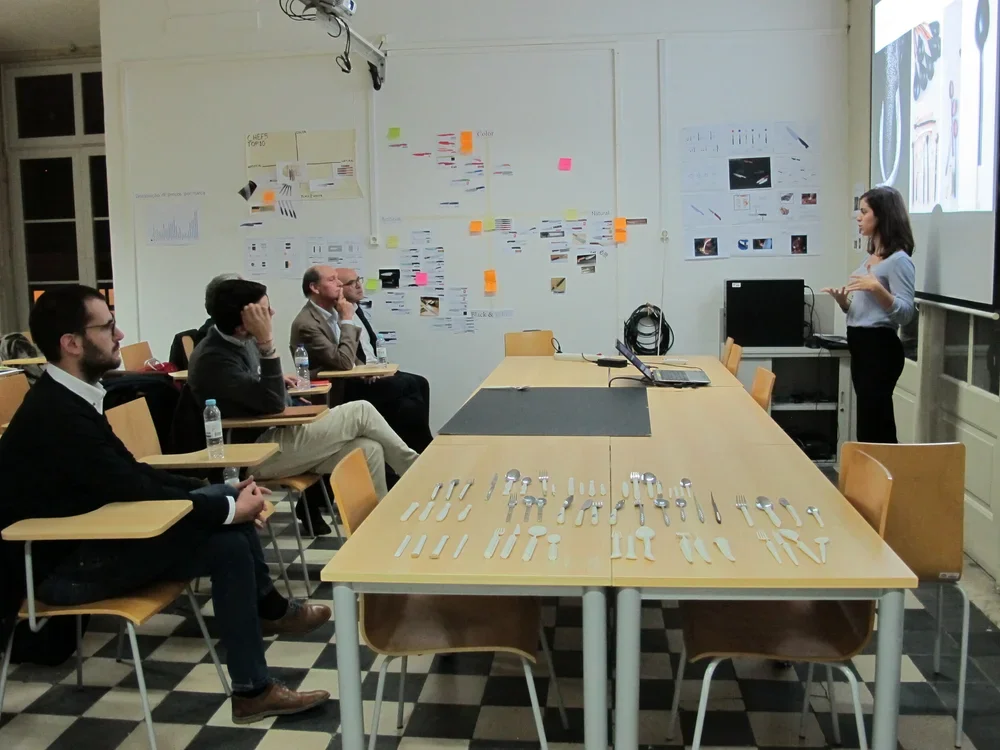 A woman is giving a presentation in a classroom with a whiteboard, while four men are seated at desks watching her. The desks have utensils laid out on them, and there are water bottles beside the seated men. The classroom has checkered flooring, and