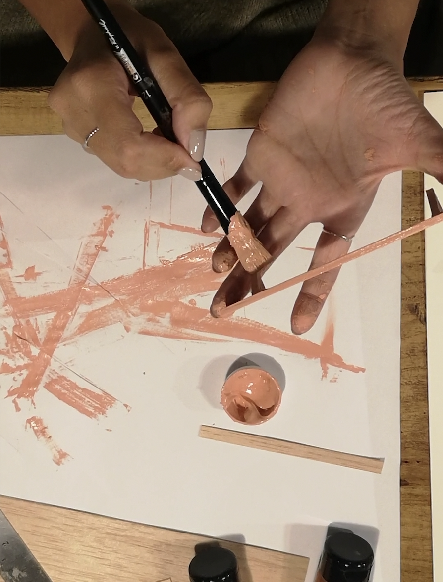 Person painting with peach-colored paint on paper, using a paintbrush and a stick, with paint on fingers and table.