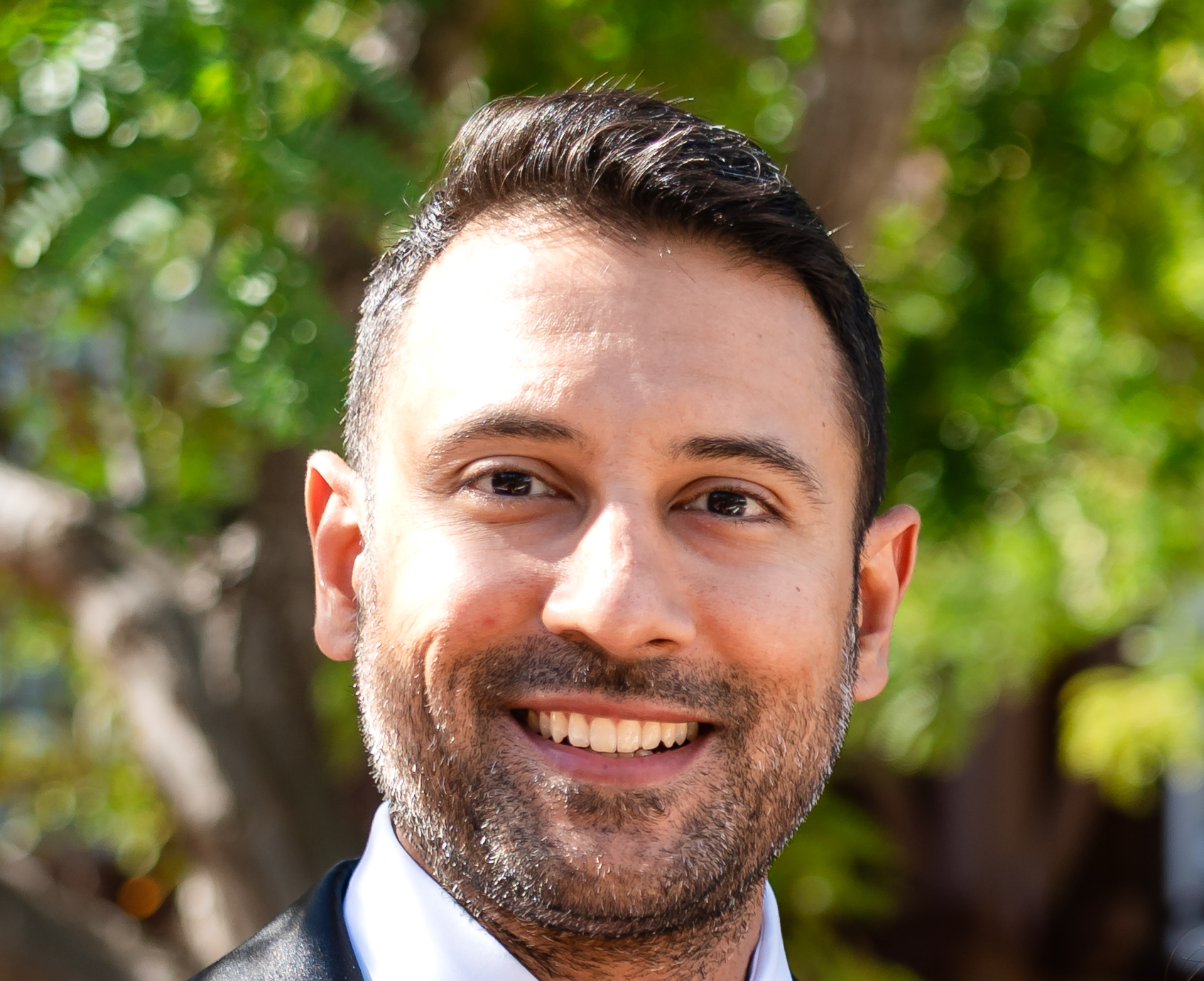 A smiling man with dark hair and a beard, wearing a suit and white shirt, standing outdoors with a background of greenery.