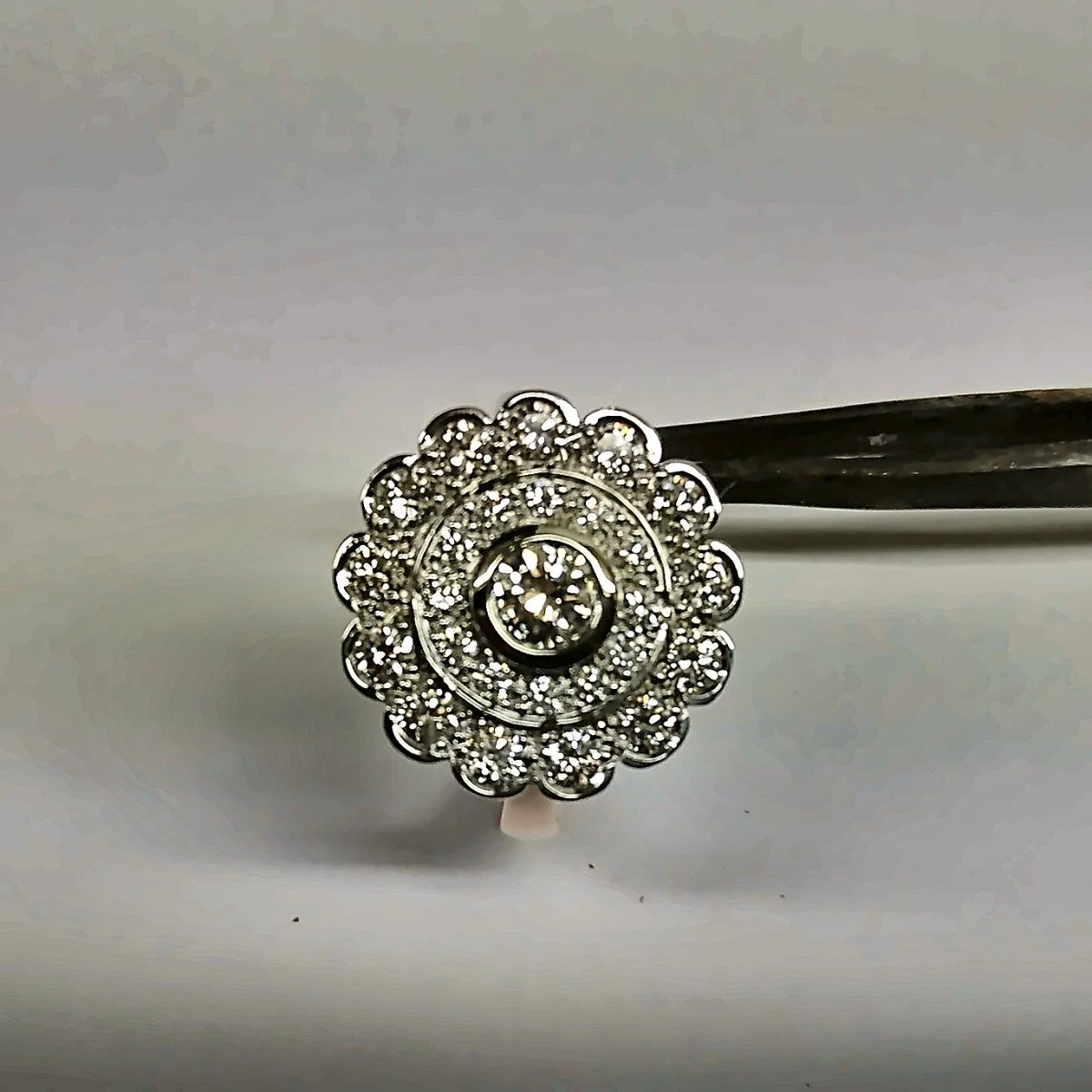 Close-up of a silver ring with a spiral design and numerous small diamonds, held by tweezers against a light background.