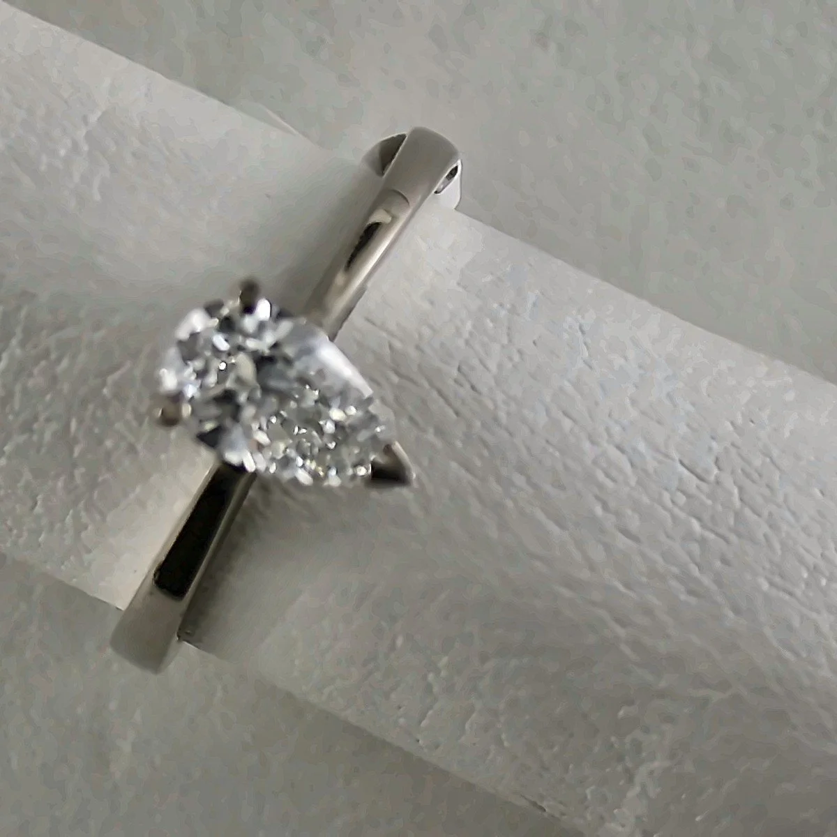 A close-up of a white gold ring with a large, diamond, attached to a white surface with a metal clamp.