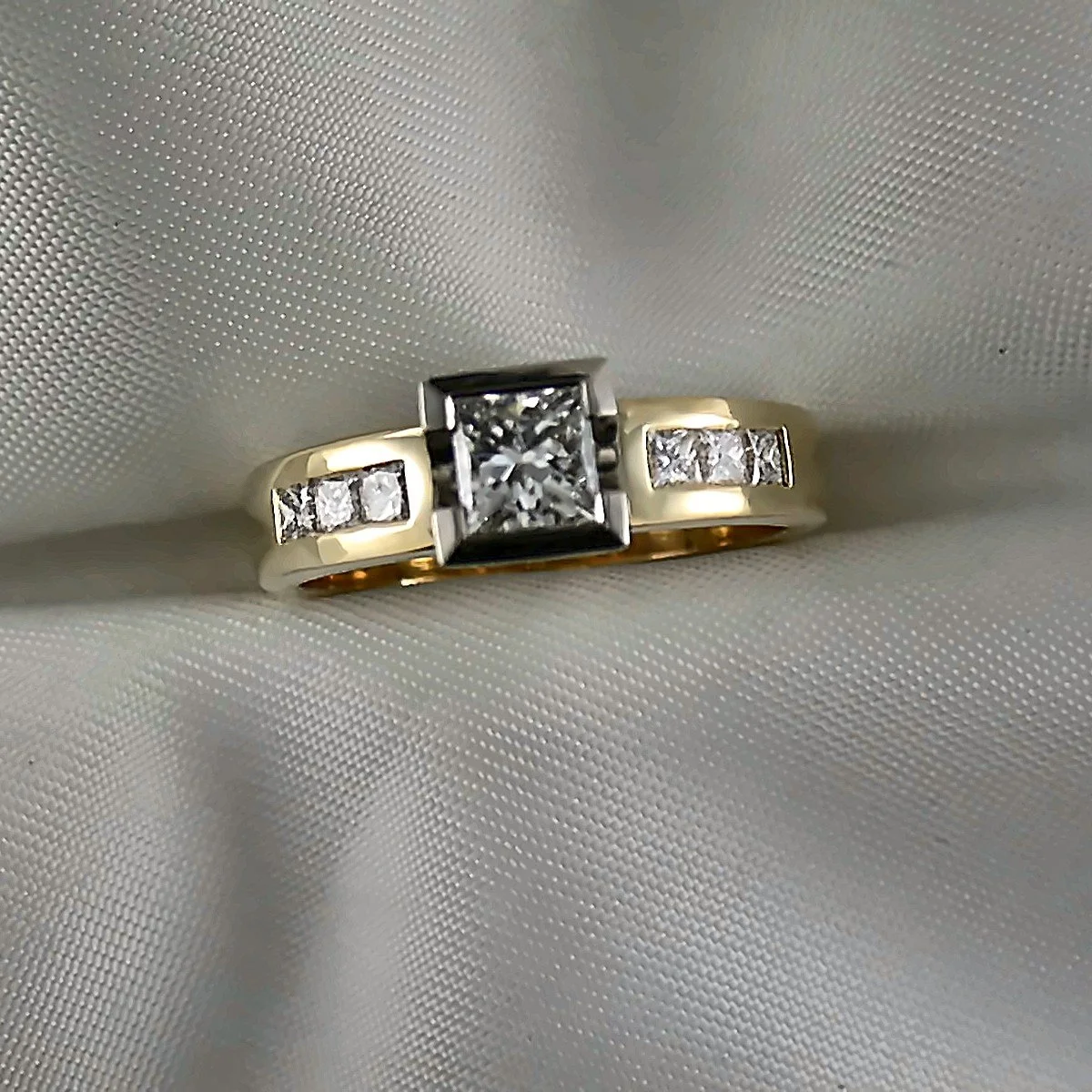 Gold ring with a square center diamond and two row of small diamonds on each side, resting on white fabric.