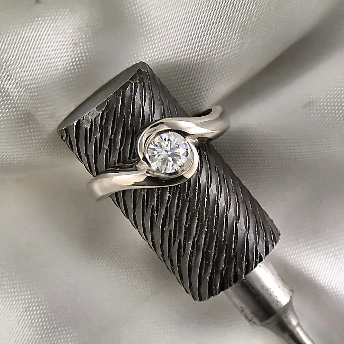 A close-up of a two-tone ring with a large round diamond set in a sleek, curved band, resting on a textured black display stand with a white fabric background.