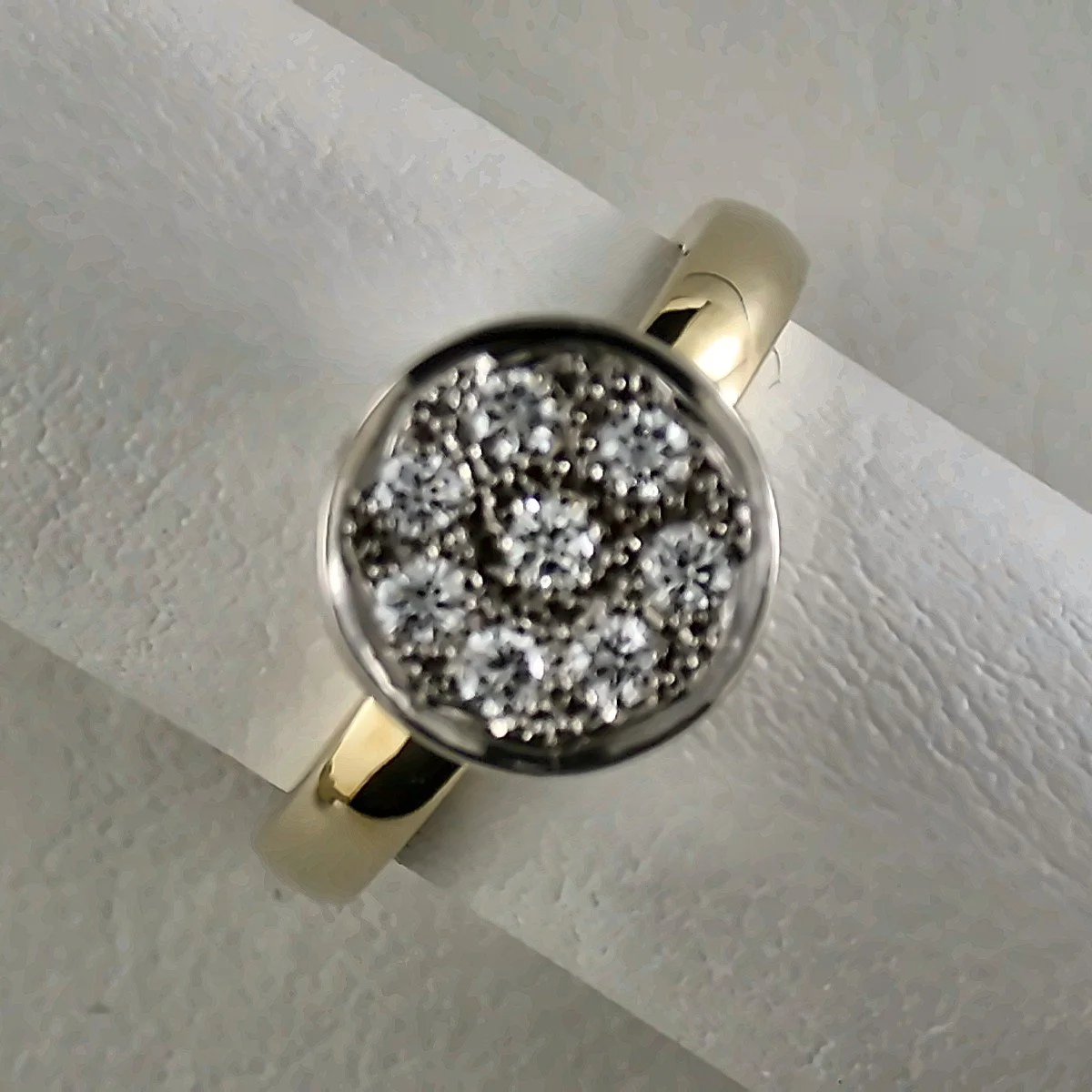Close-up of a gold ring with a large round face filled with black, white, and silver small beads or stones, placed on a white textured paper surface.