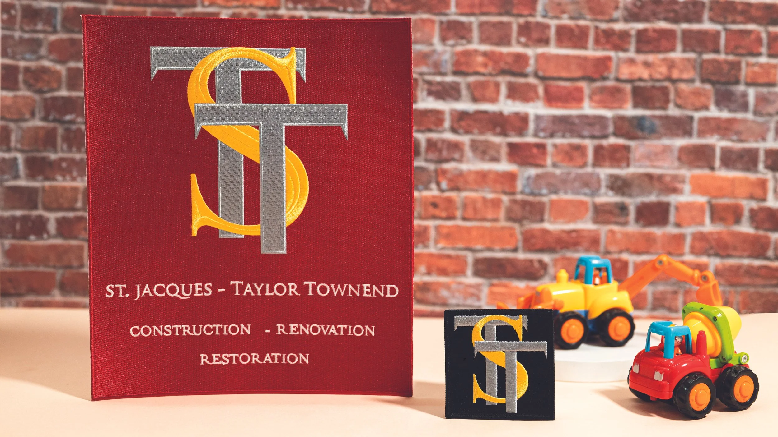 Large and small embroidered logo patches for St. Jacques Taylor Townend construction, shown against a brick wall

