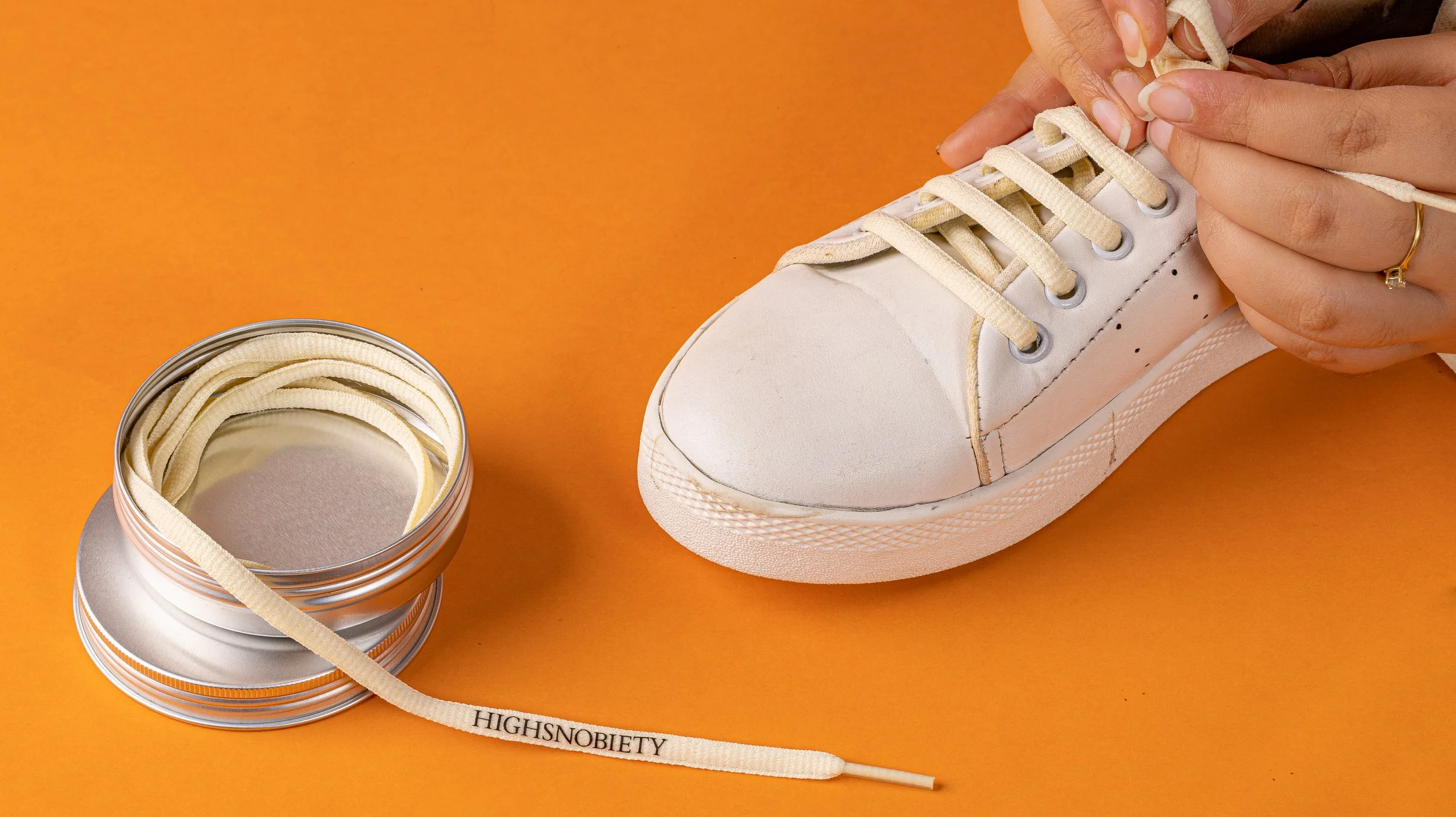 Cream printed shoelaces being laced into a white sneaker, with a round metal presentation tin
